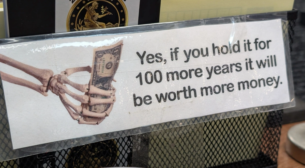 Coin shop sign showing skeleton hands holding a dollar bill with the message "If you hold it for 100 more years it will be worth more money"